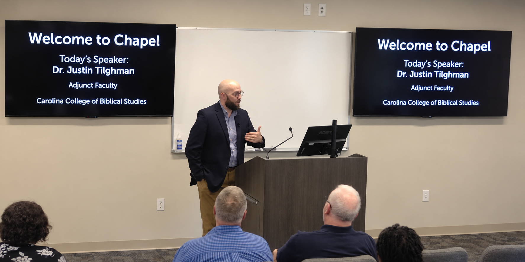 Justin Tilghman speaking beside a lectern to a seated chapel audience at Carolina College of Biblical Studies.